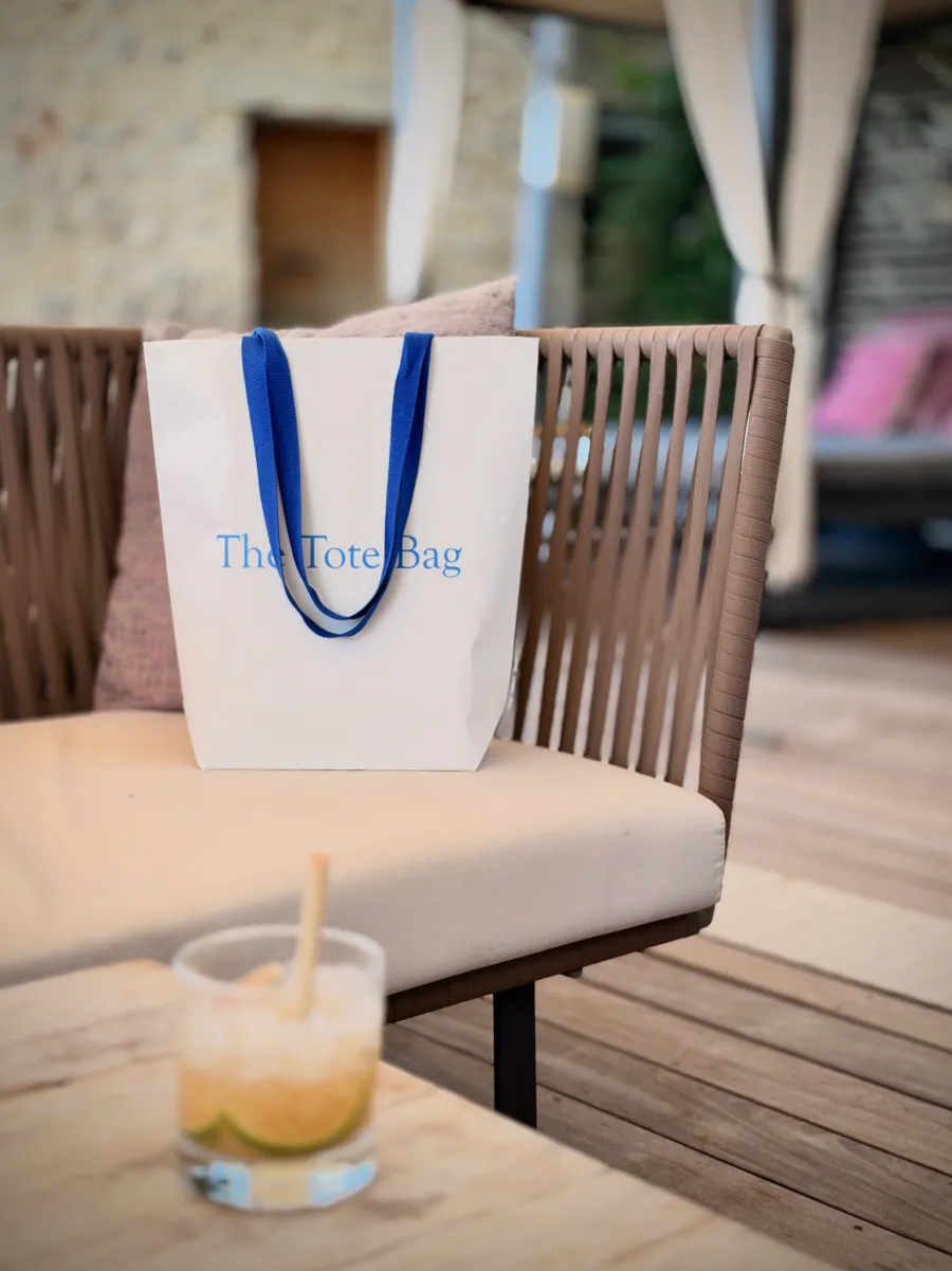 The Tote Bag posé sur un fauteuil de terrasse, à côté d'un cocktail